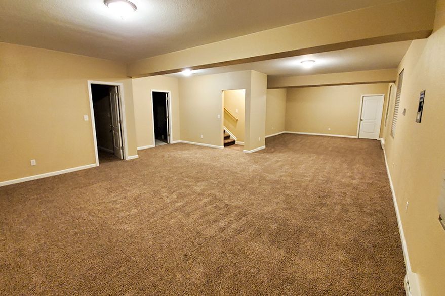 Empty beige-walled room with brown carpet, doorways, and a staircase.