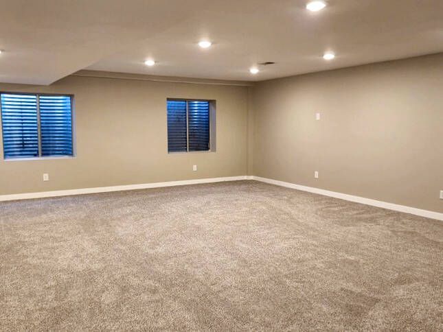 Empty beige-walled basement room with gray carpet and two small windows; several recessed lights.