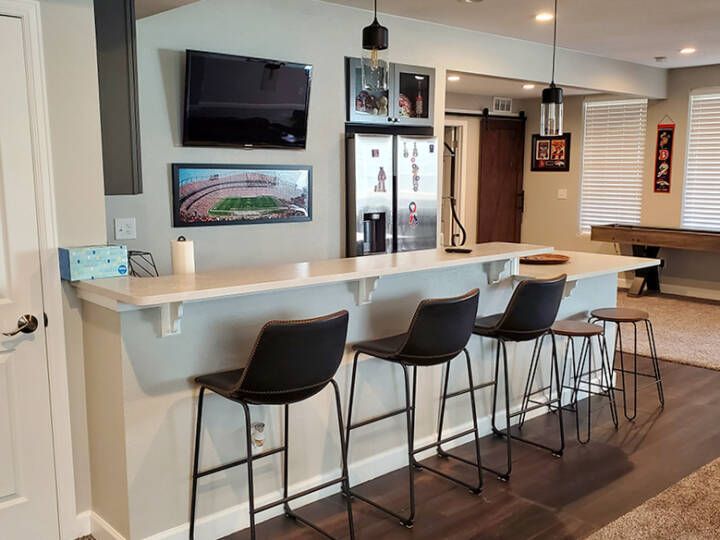Basement bar with bar stools, refrigerator, TVs, pendant lights, and a doorway to a game room.