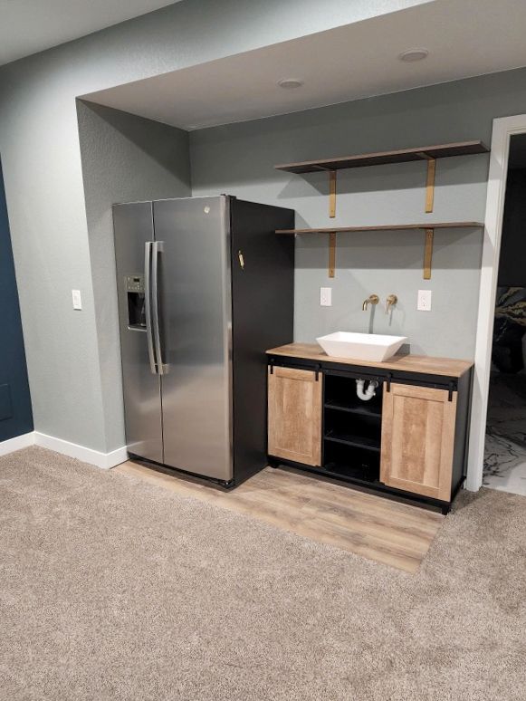 A stainless steel refrigerator beside a small bar with a sink and shelving. Light gray walls, carpet.