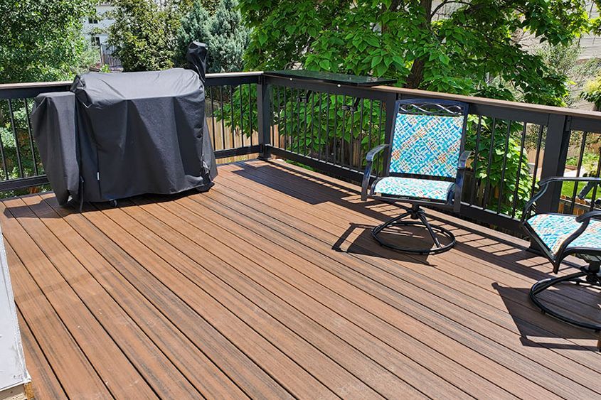 Wooden deck with grill covered by black tarp, two chairs, and a railing with trees in the background.