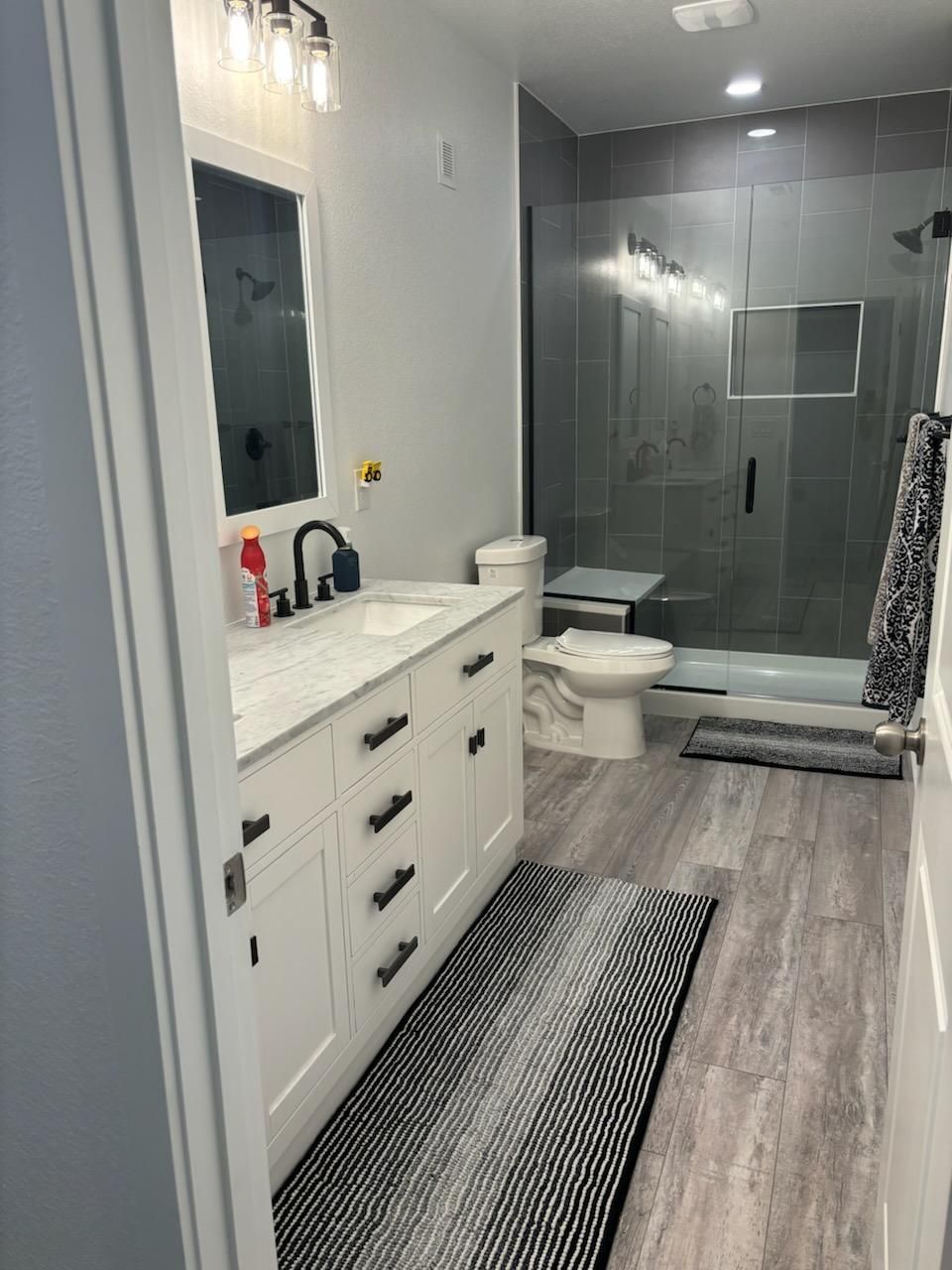 White bathroom with gray shower, toilet, vanity, and wooden floor. Black accents and a long rug.