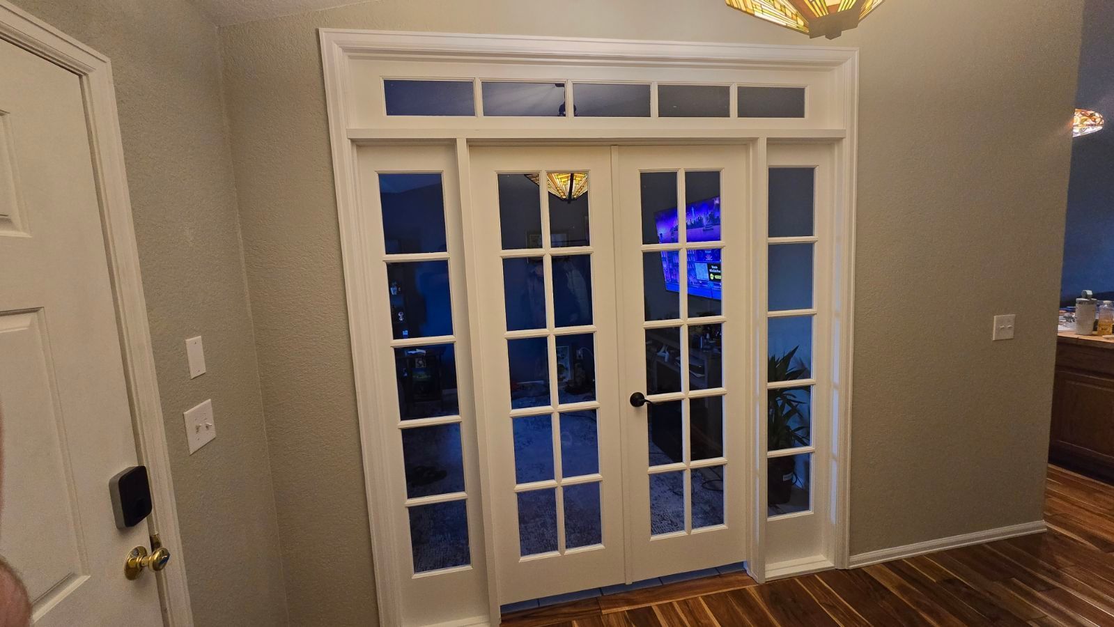 White french doors with grid pattern, flanked by sidelights, opening to a brightly lit area.