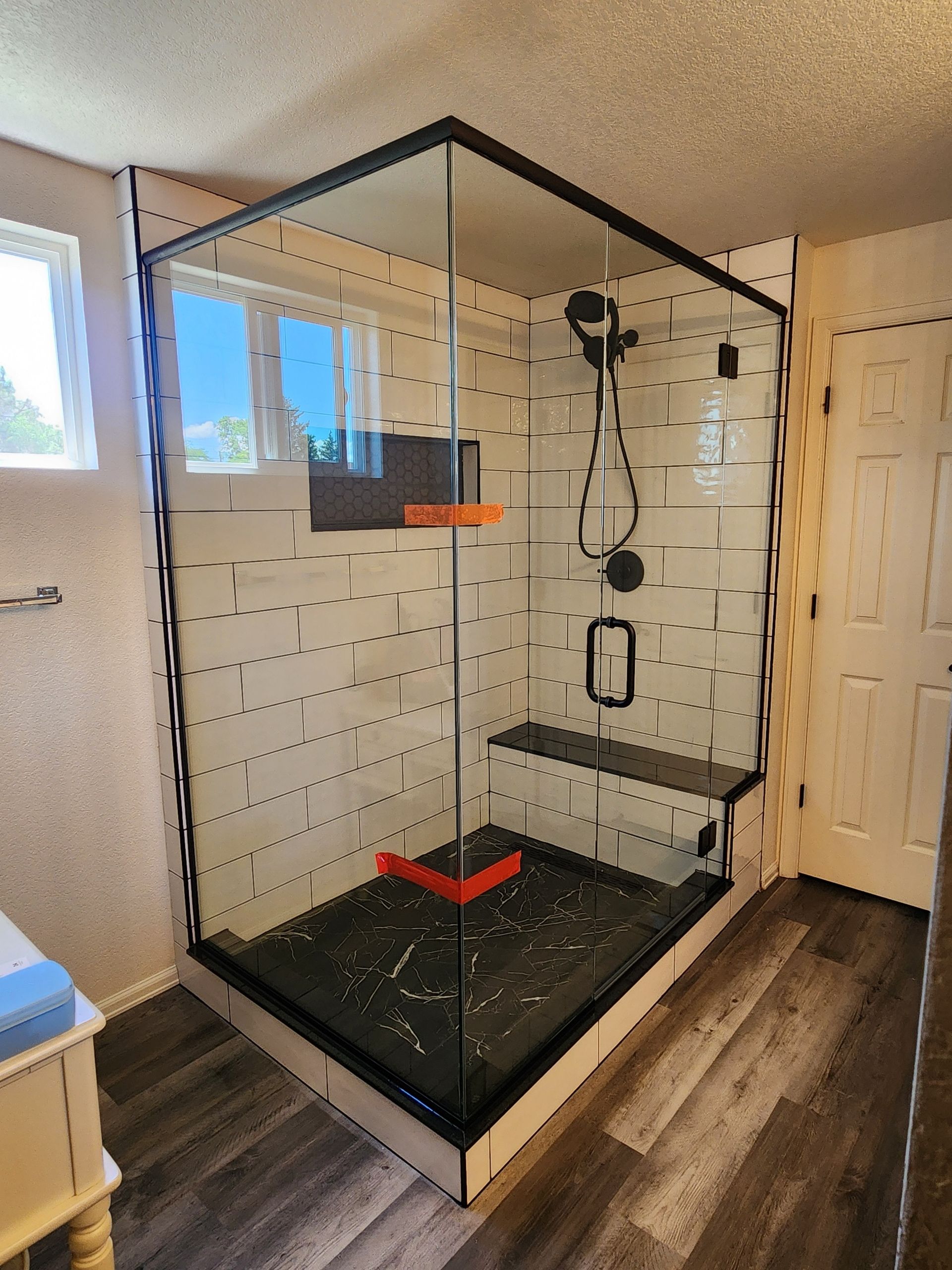 Glass shower with black trim, seat, and fixtures; white tiled walls and dark floor.
