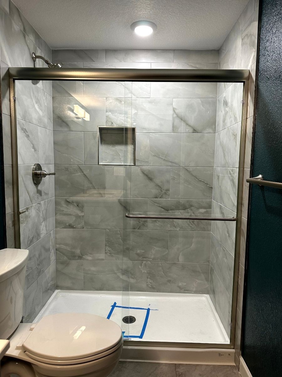Shower with marble-look tiles, glass door, and bronze frame, toilet on left.