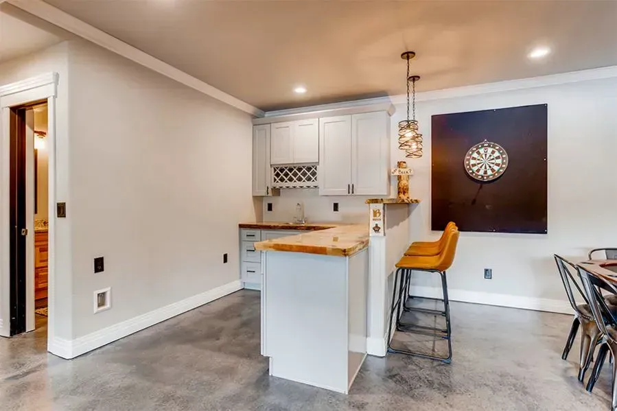 A gray bar with a dartboard, stools, cabinets, and open doorway in a room with polished concrete flooring.