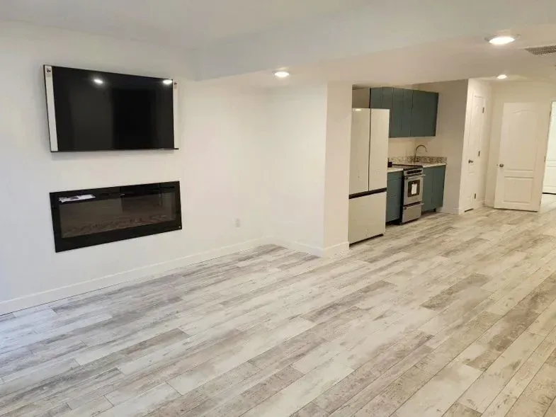 Living room with a mounted TV, fireplace, and small kitchen with teal cabinets. Light wood flooring.