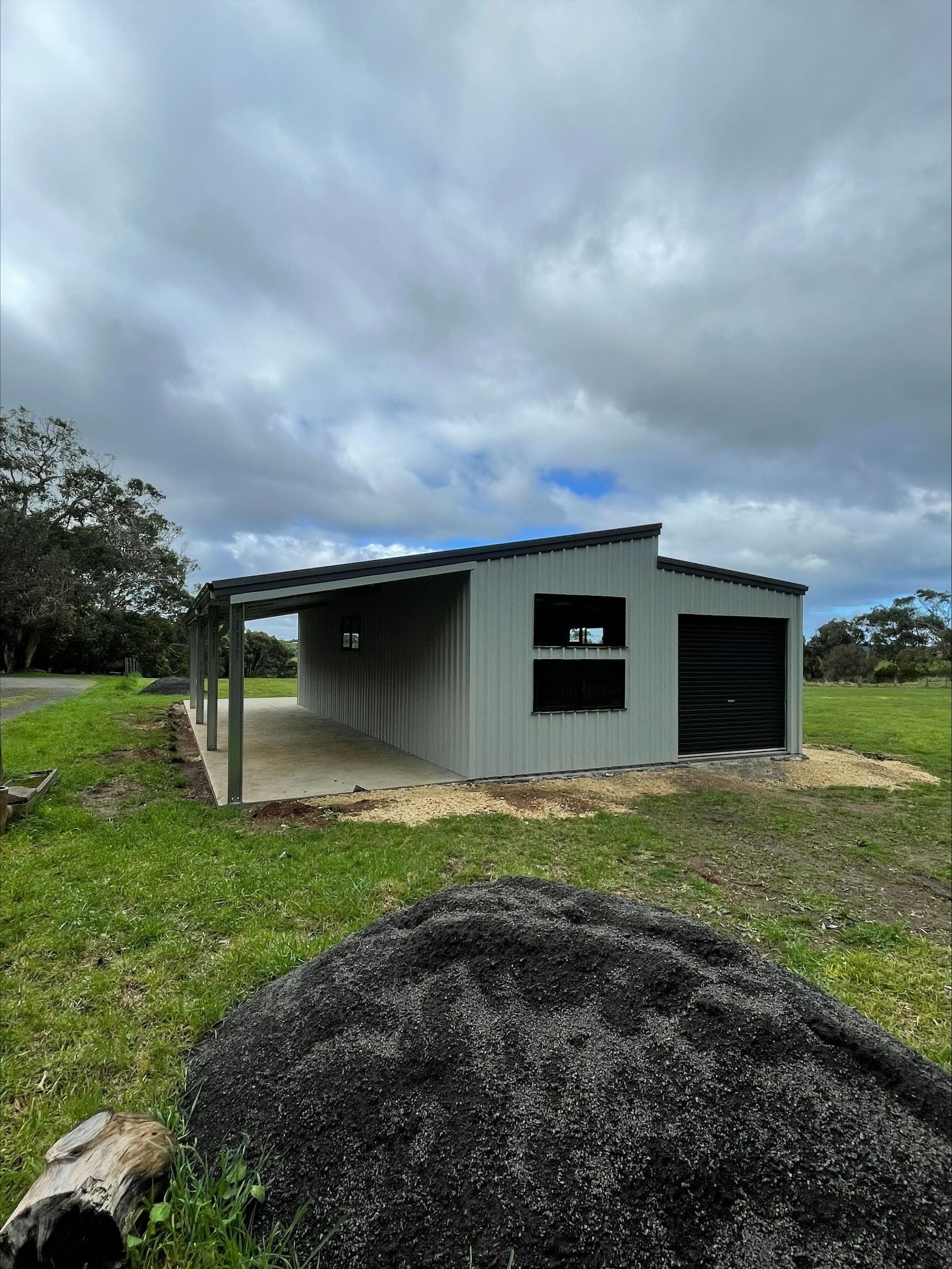 Metal shed with a carport, set in a grassy field under a cloudy sky.