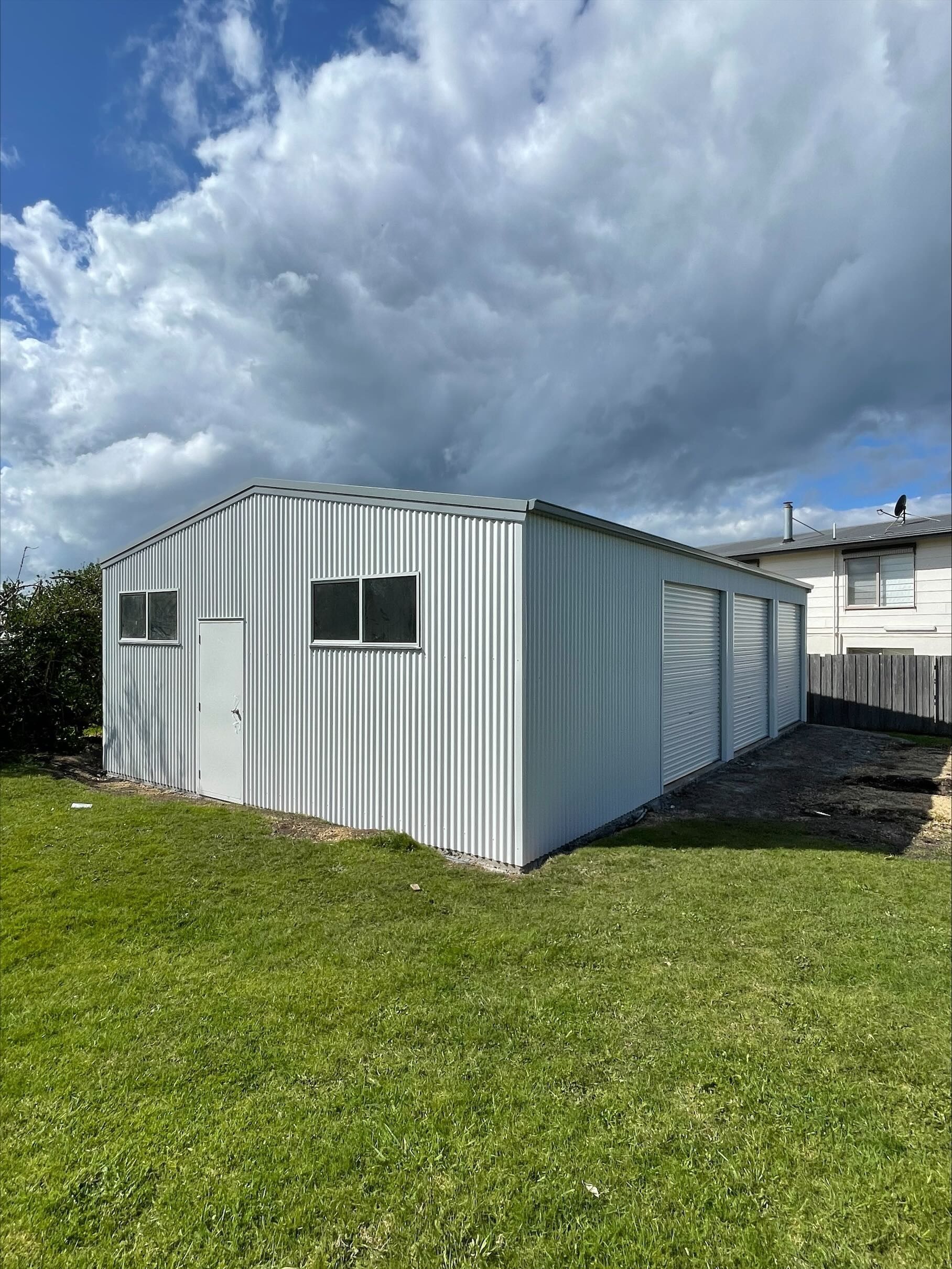 White metal shed with a gabled roof, two small windows, and two large garage doors, set on green grass under a cloudy sky.