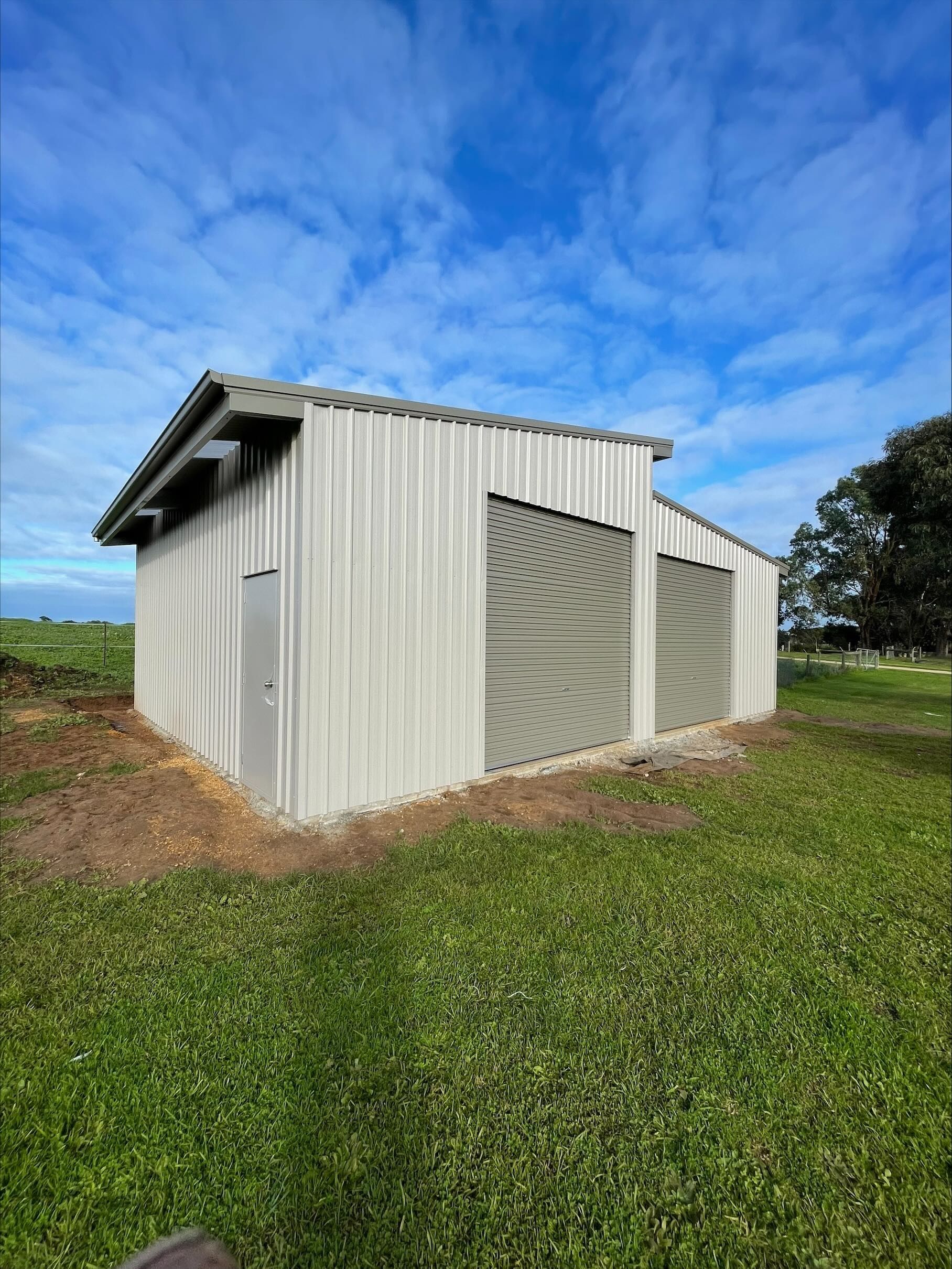 A light gray shed with two screened doors stands in a green field under a cloudy blue sky.