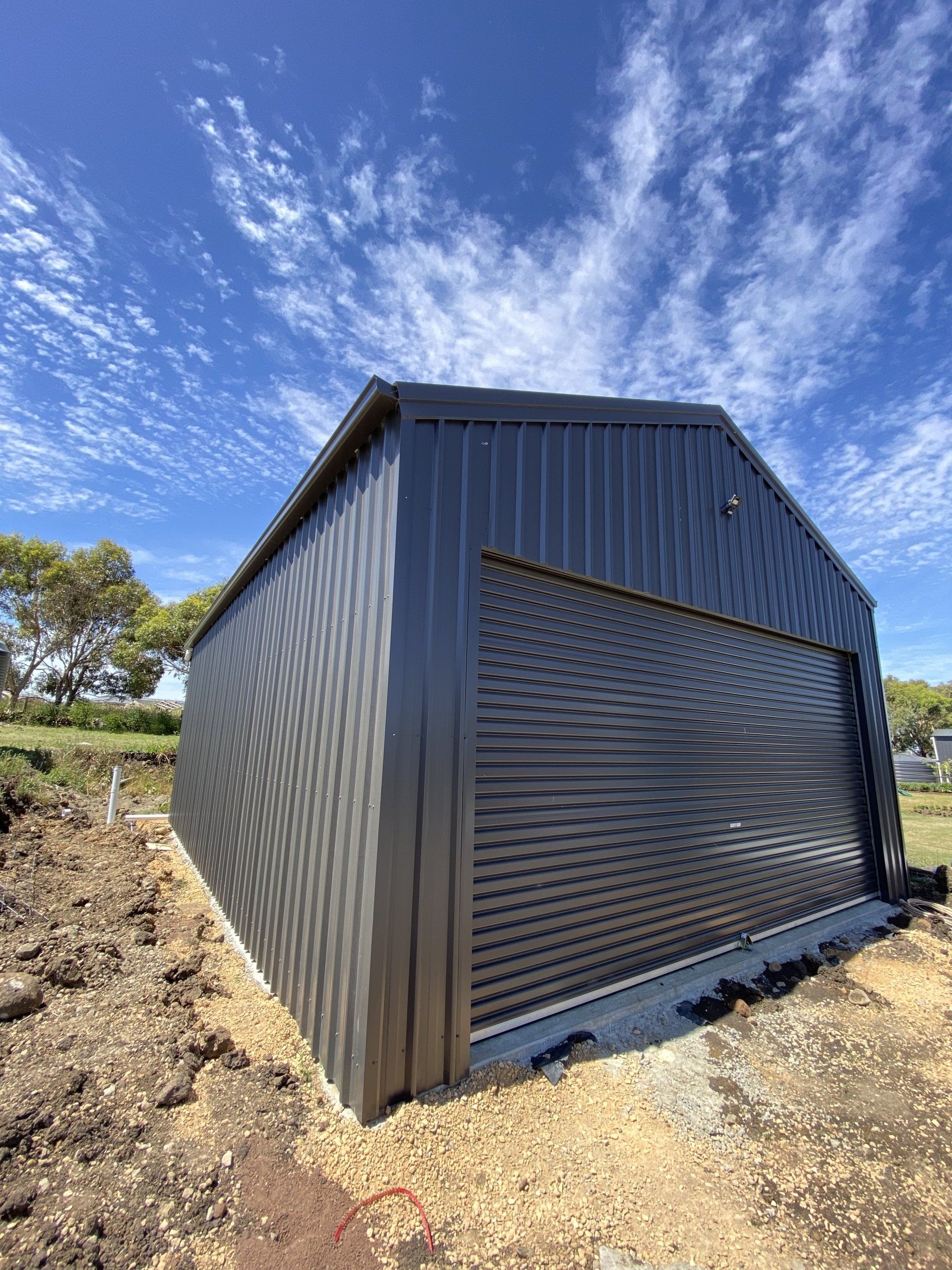 Dark gray metal shed with a roller door under a blue sky with clouds.