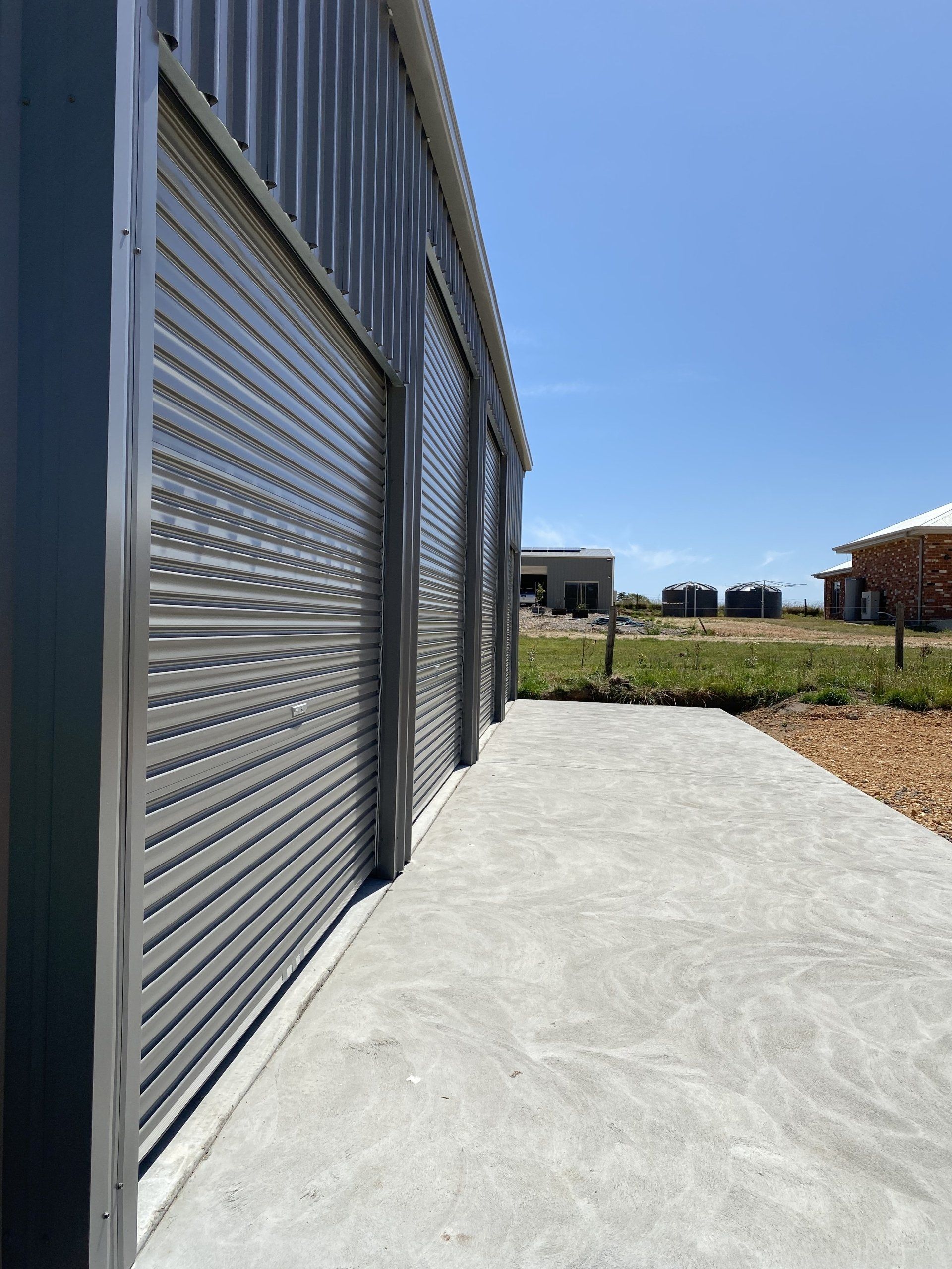 Gray metal building with three closed roll-up doors, concrete path, and blue sky.