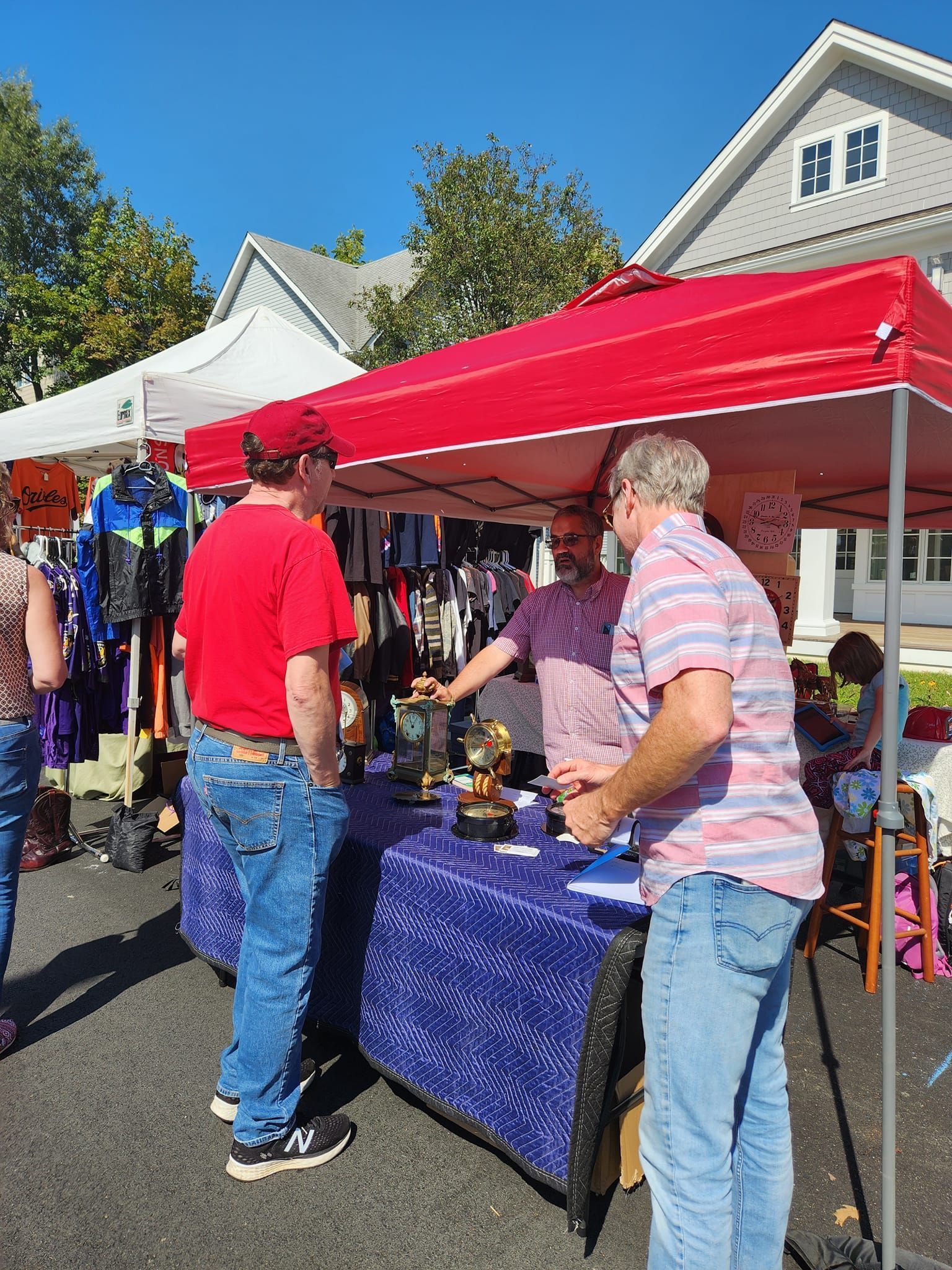 Three people stand under a red tent at an outdoor market, looking at items on a table draped with a patterned blue cloth.