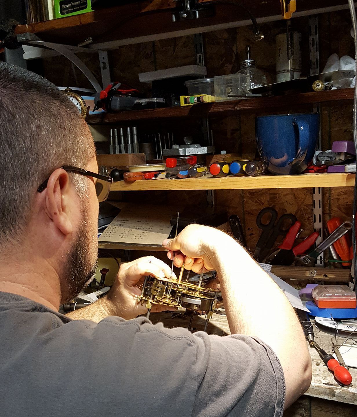 A person focuses intently on repairing a small, metallic clock mechanism at a cluttered workbench with various tools.