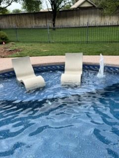 Two beige chaise lounge chairs in a pool, near a fountain. Green grass and a wooden fence in the background.