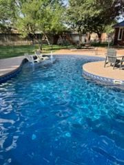 Swimming pool with blue water, surrounded by concrete. Chairs sit on the pool's edge, trees in background.