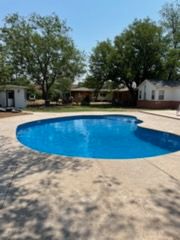 Blue swimming pool surrounded by concrete patio, with trees and buildings in the background on a sunny day.