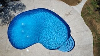 Aerial view of a kidney-shaped swimming pool with blue water and surrounding concrete patio.