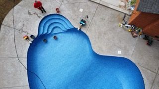 Overhead view of a bright blue, kidney-shaped pool under construction, with workers around the edges.