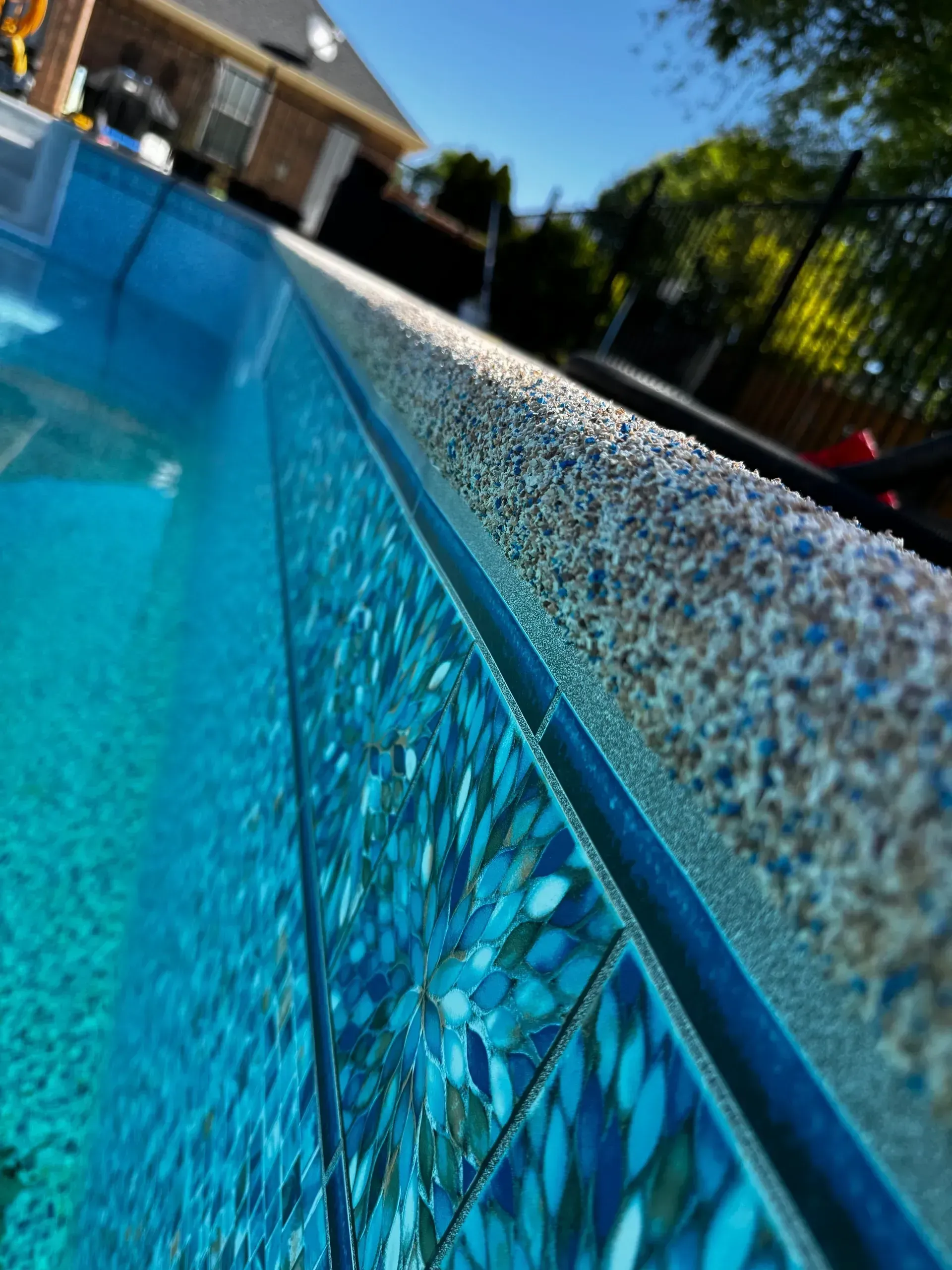 Close-up of a pool's edge, showing blue mosaic tile and textured coping with clear, blue water.