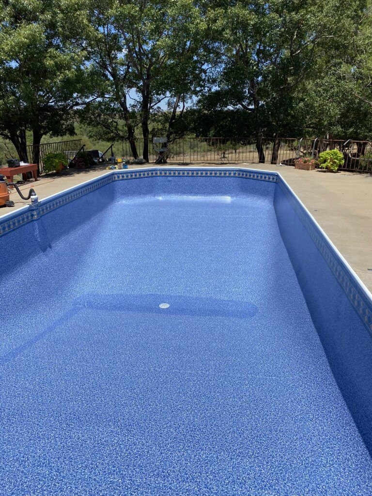 Empty swimming pool with blue tiled interior. Concrete surround and trees in background on a sunny day.