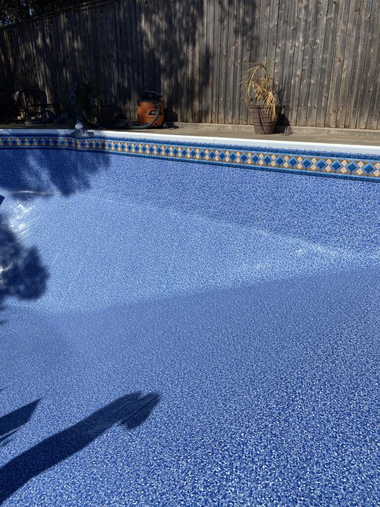 Blue mosaic-tiled swimming pool with a wooden fence in the background, on a sunny day.