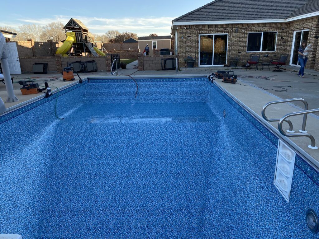 Empty, blue-lined swimming pool with handrails, surrounded by concrete patio and a brick house. Tools and playground visible.