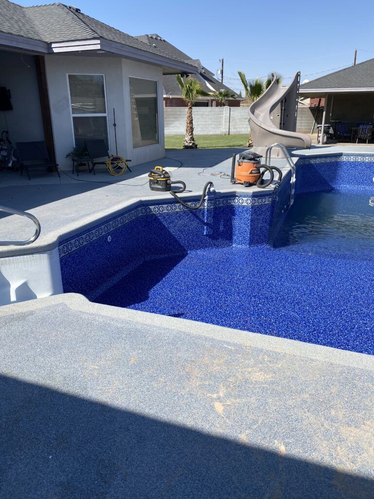 Swimming pool with blue tile, concrete patio, and a backyard with a house and slide.