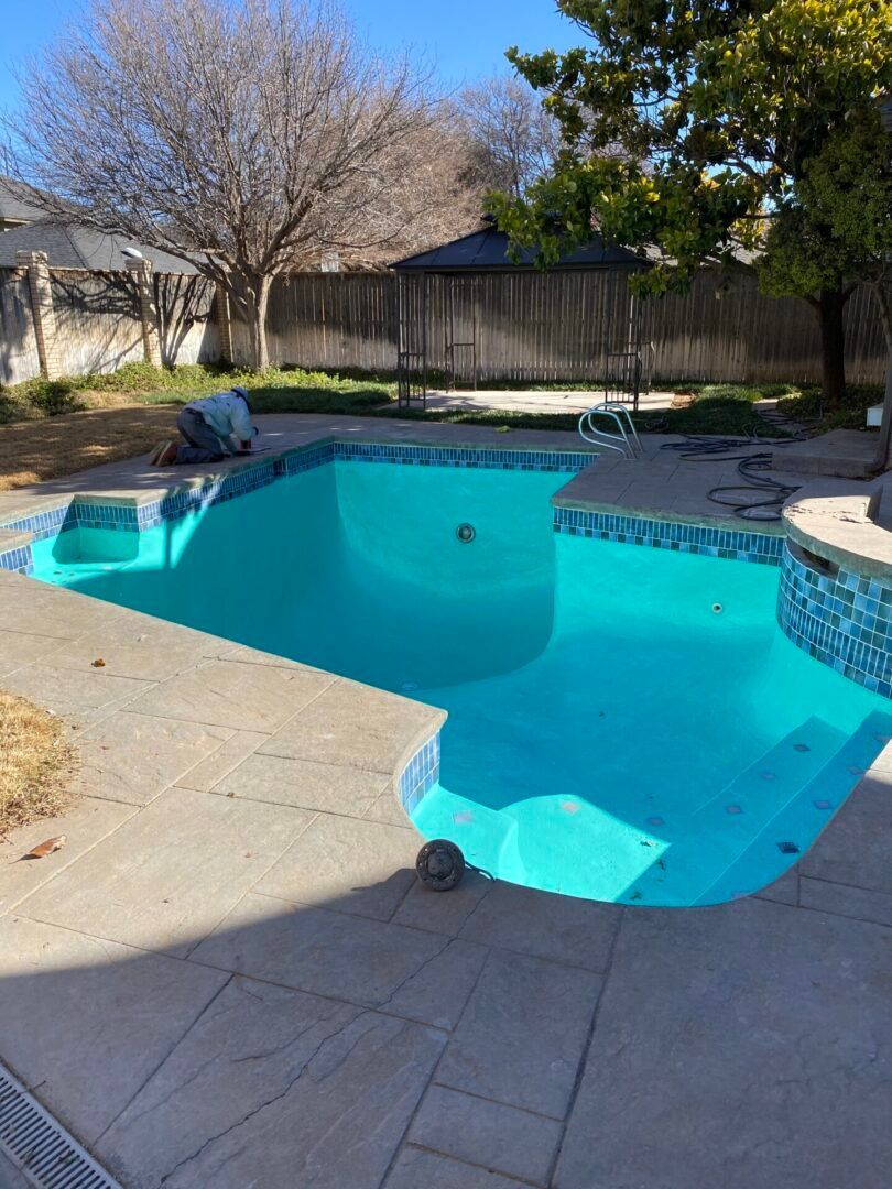 A turquoise pool surrounded by concrete, in a backyard. Trees and a fence are in the background.
