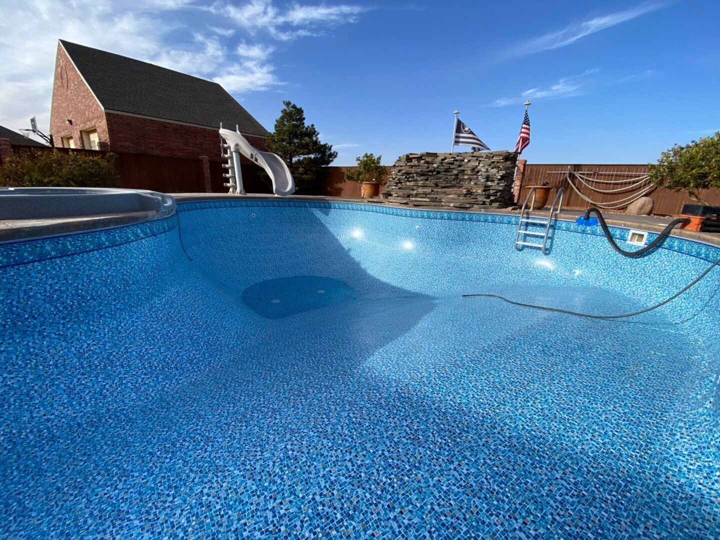 Empty blue swimming pool with slide, flag, and brick building under a sunny sky.