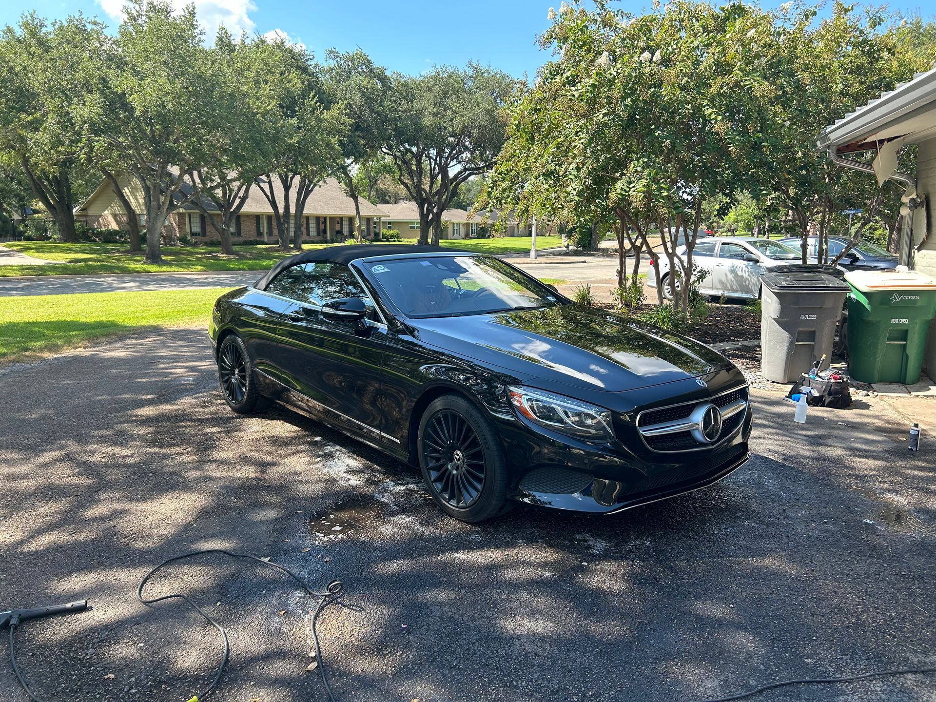 Black Mercedes convertible parked on a driveway in front of a house, sunny day.