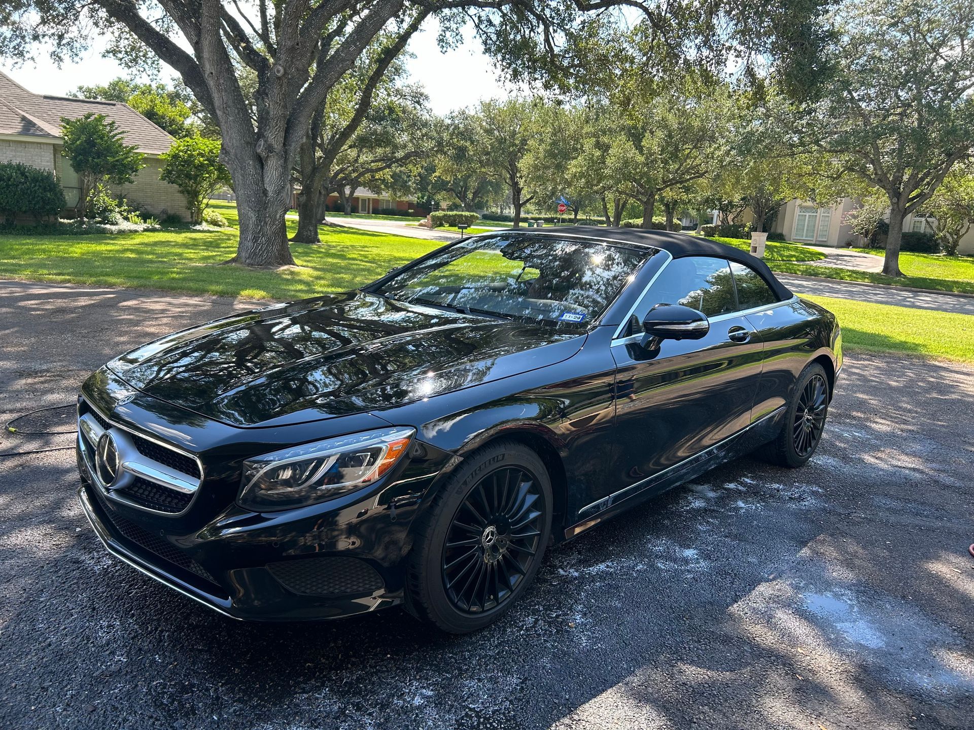 Black Mercedes coupe parked on a driveway in a suburban setting with trees and a house.