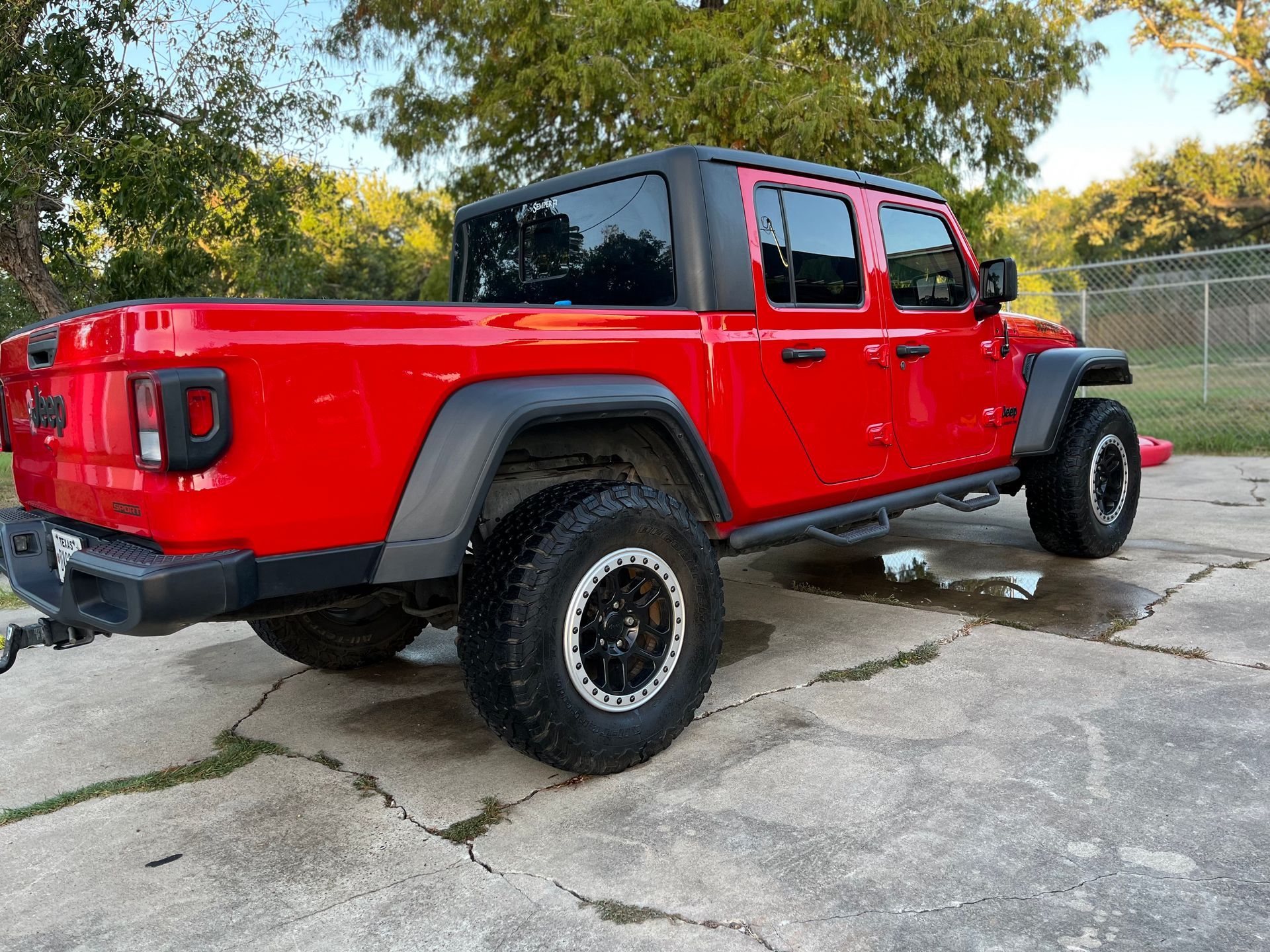 Red Jeep Gladiator truck on a concrete surface, black accents, large tires, and a black hard top.
