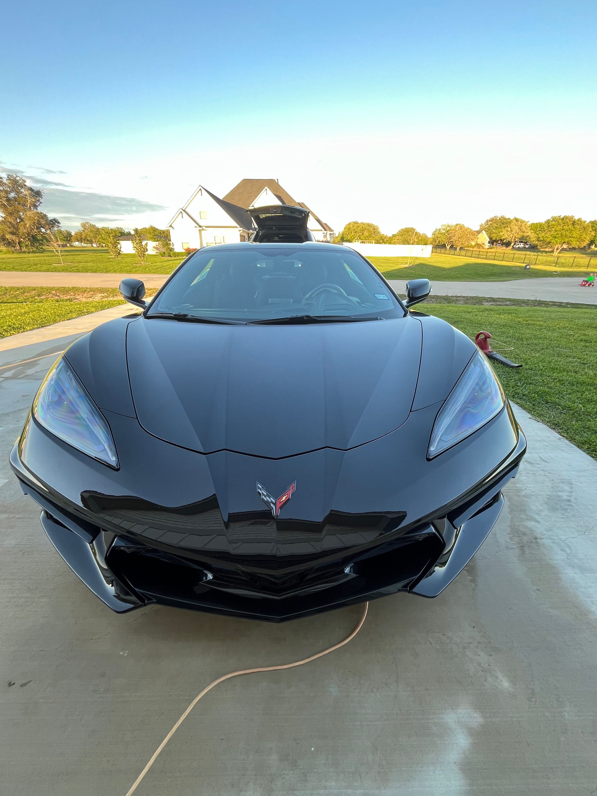 Black Corvette sports car parked on a concrete driveway with a house and trees in the background.