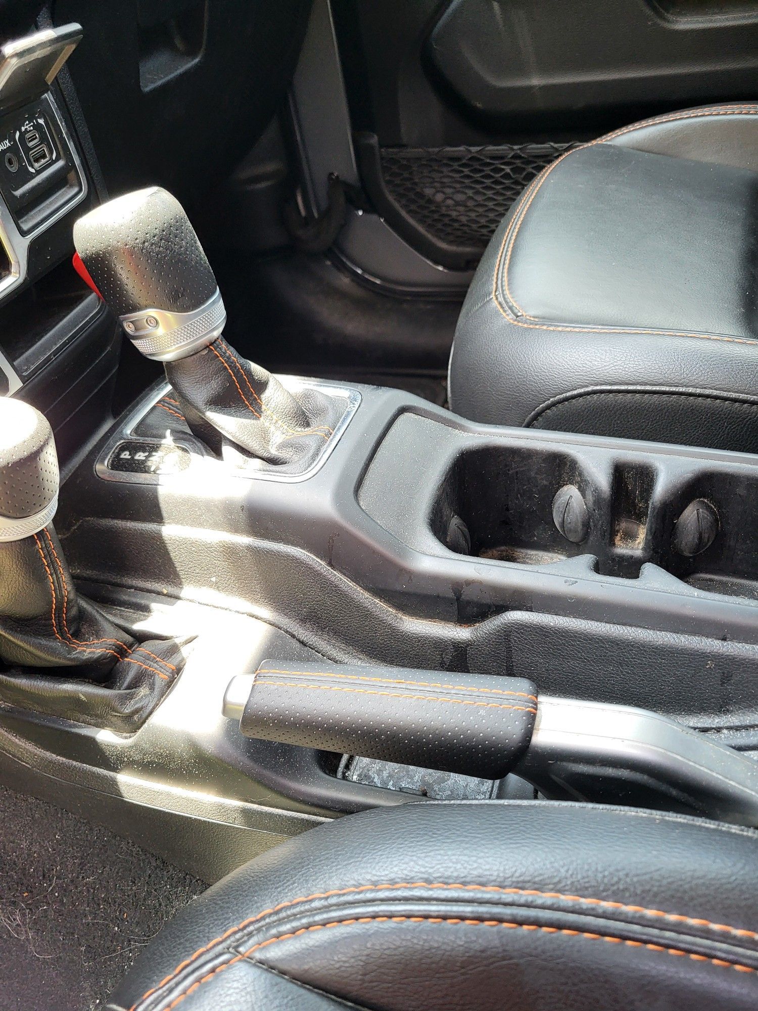 Interior of a Jeep Wrangler: gear shift, cup holders, parking brake, and black leather seats with orange stitching.