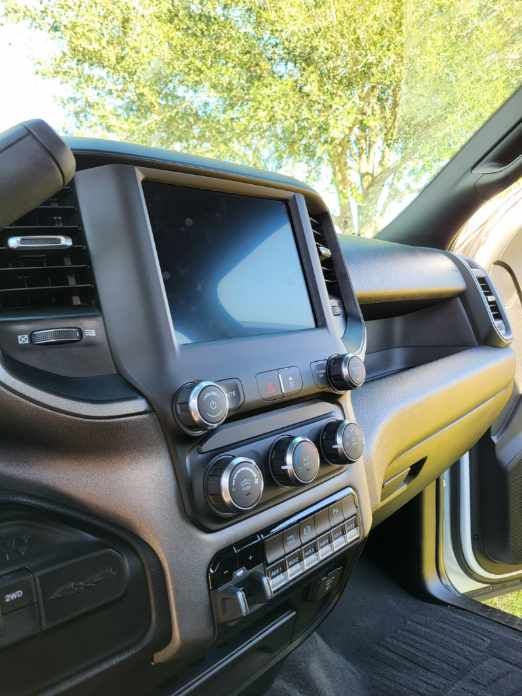 Interior of a Ram truck dashboard: large touchscreen, knobs, and buttons. Brown and black with green trees in background.