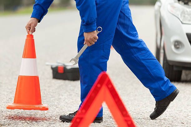 Mechanic in blue overalls places a safety cone and carries a wrench near a car on a road.