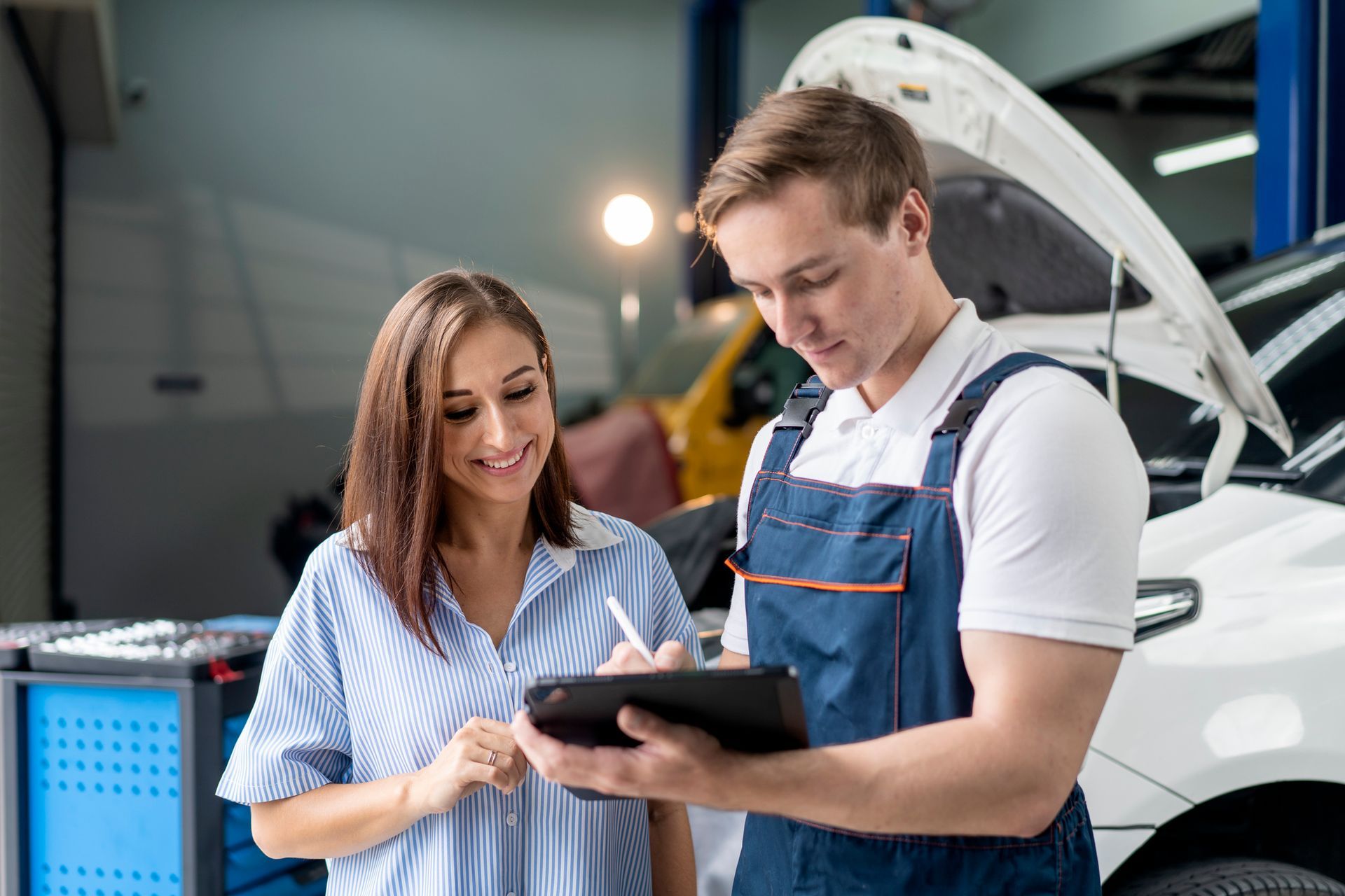 Mechanic in overalls reviews tablet with a woman in a striped shirt near a car in a repair shop.