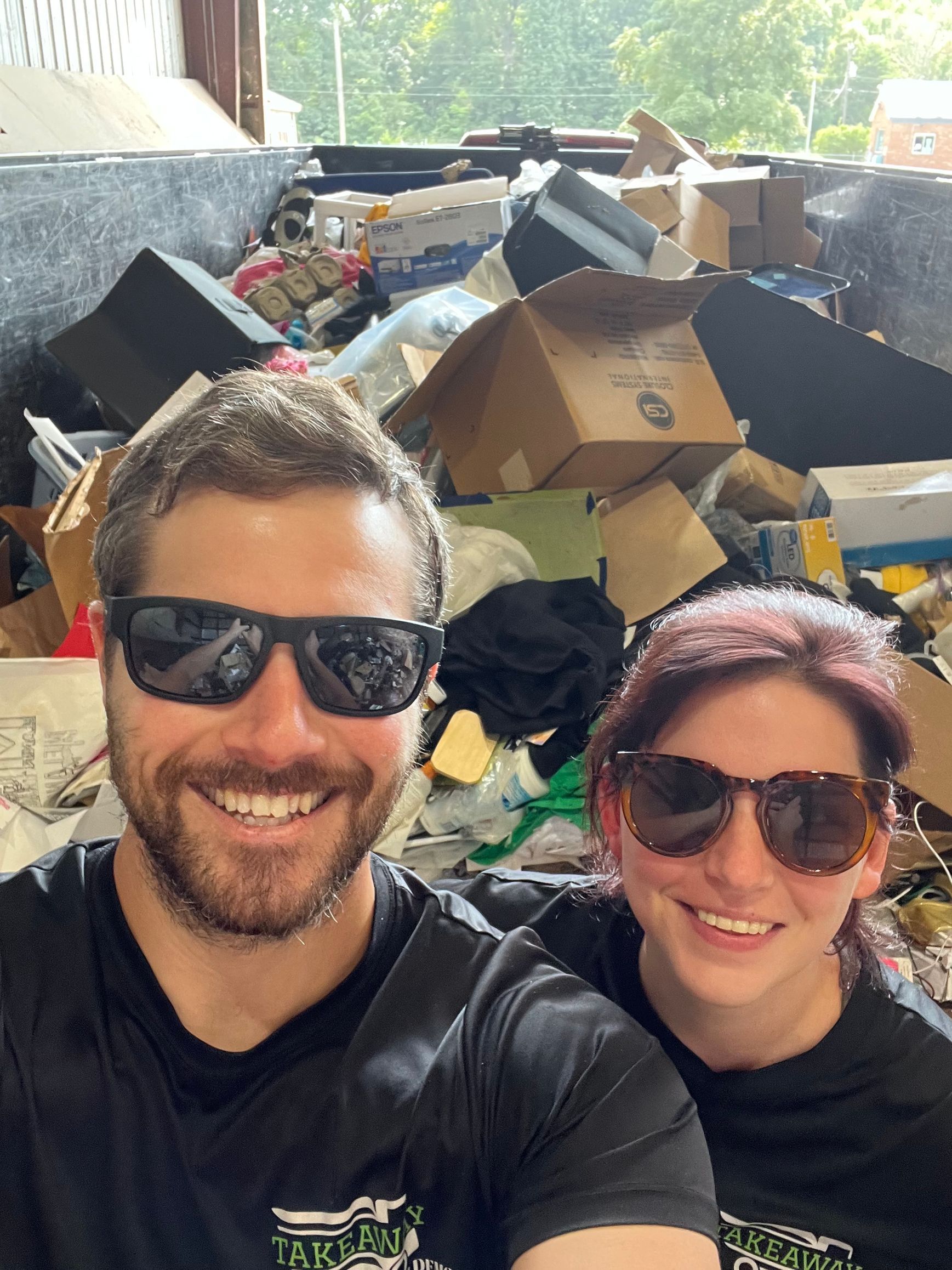 A man and a woman wearing sunglasses are posing for a picture in front of a pile of trash.