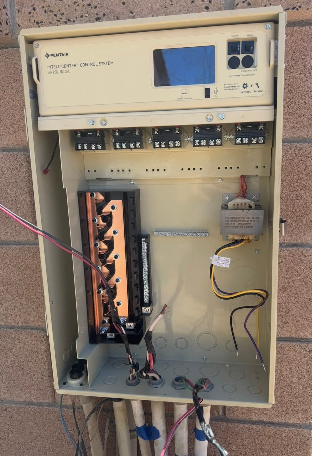 Tan electrical control panel on a brick wall with exposed wiring and a digital display.