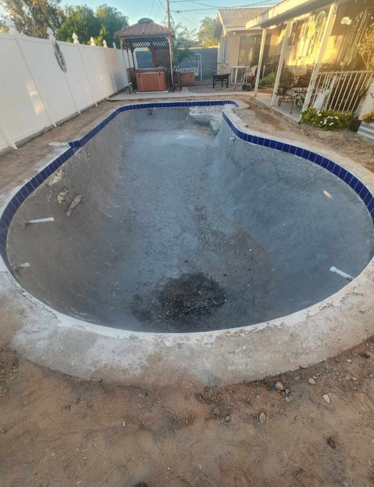 Empty, kidney-shaped swimming pool with blue tiled border, surrounded by concrete and dirt, outdoors.