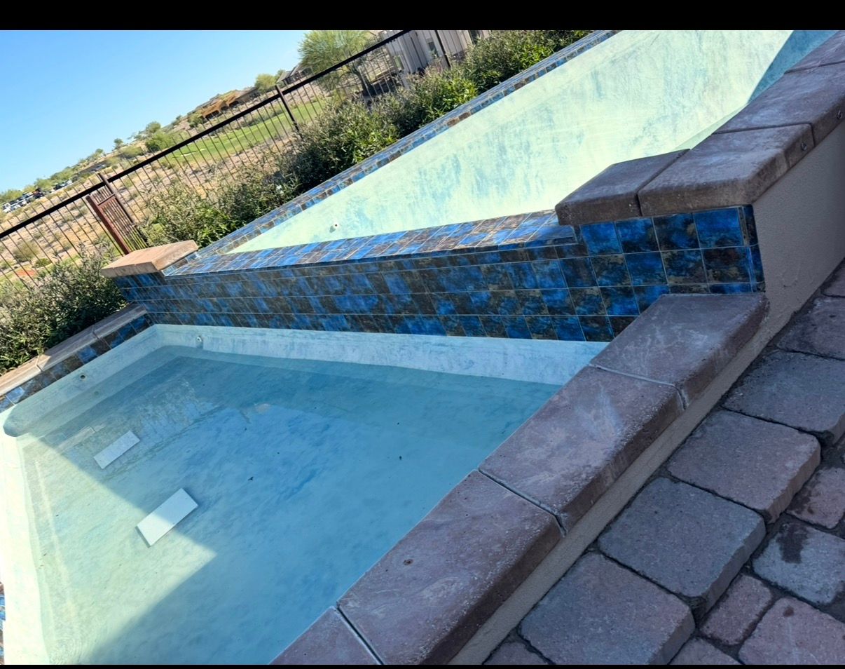 Empty swimming pool with blue-tiled waterfall feature and surrounding stone patio.