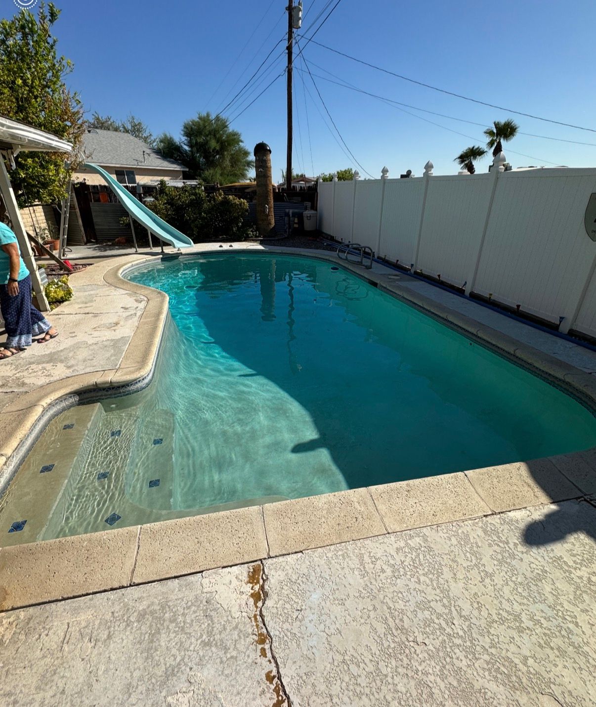 A blue swimming pool in a backyard with a slide and a white fence under a clear sky.
