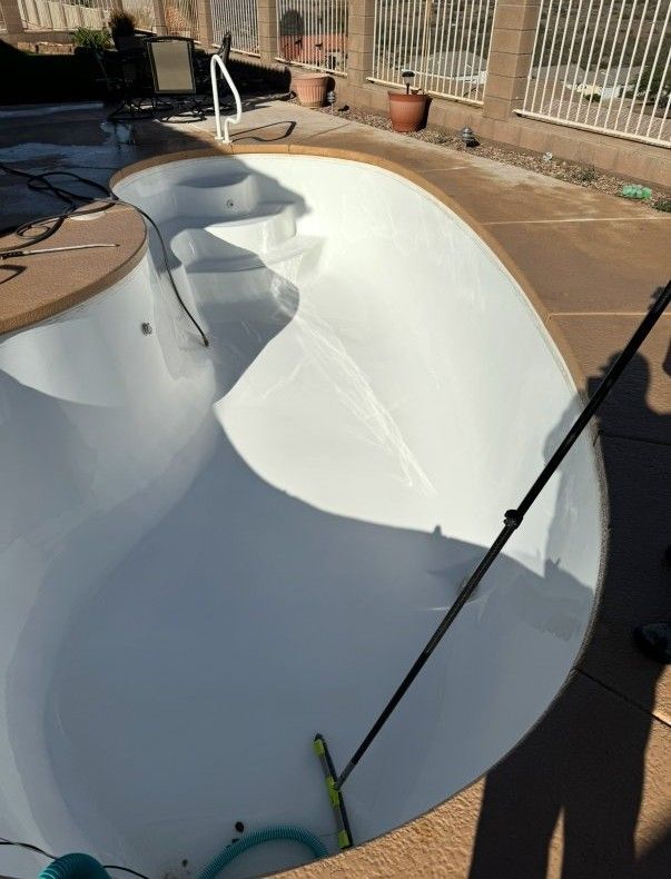 Empty white pool with brown coping and concrete deck, being cleaned with a pool brush.