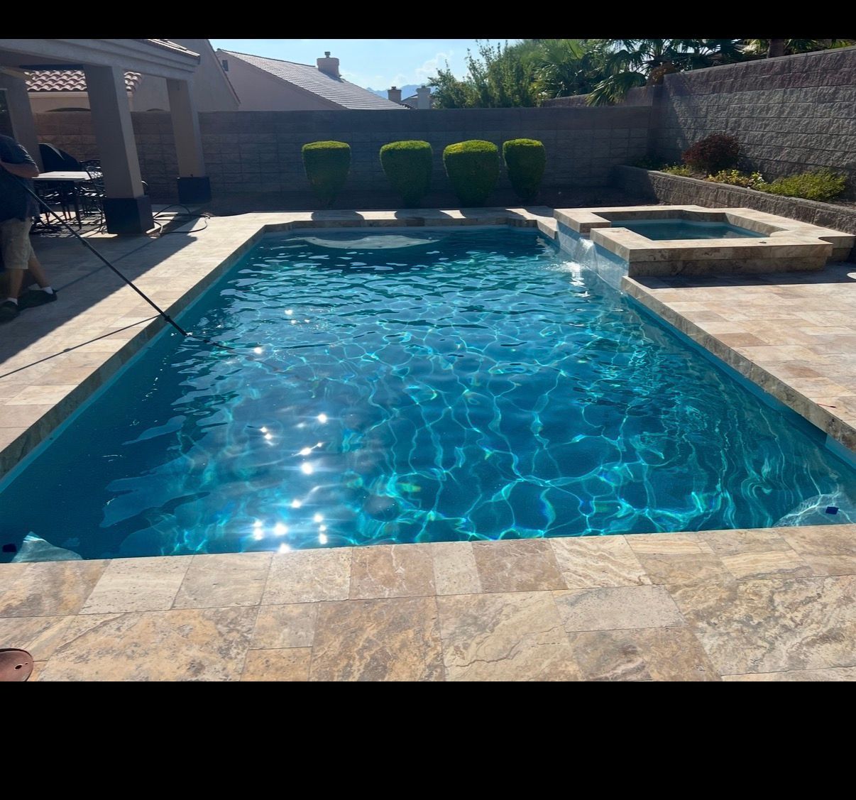 A rectangular blue swimming pool and spa surrounded by stone patio in a sunny backyard.