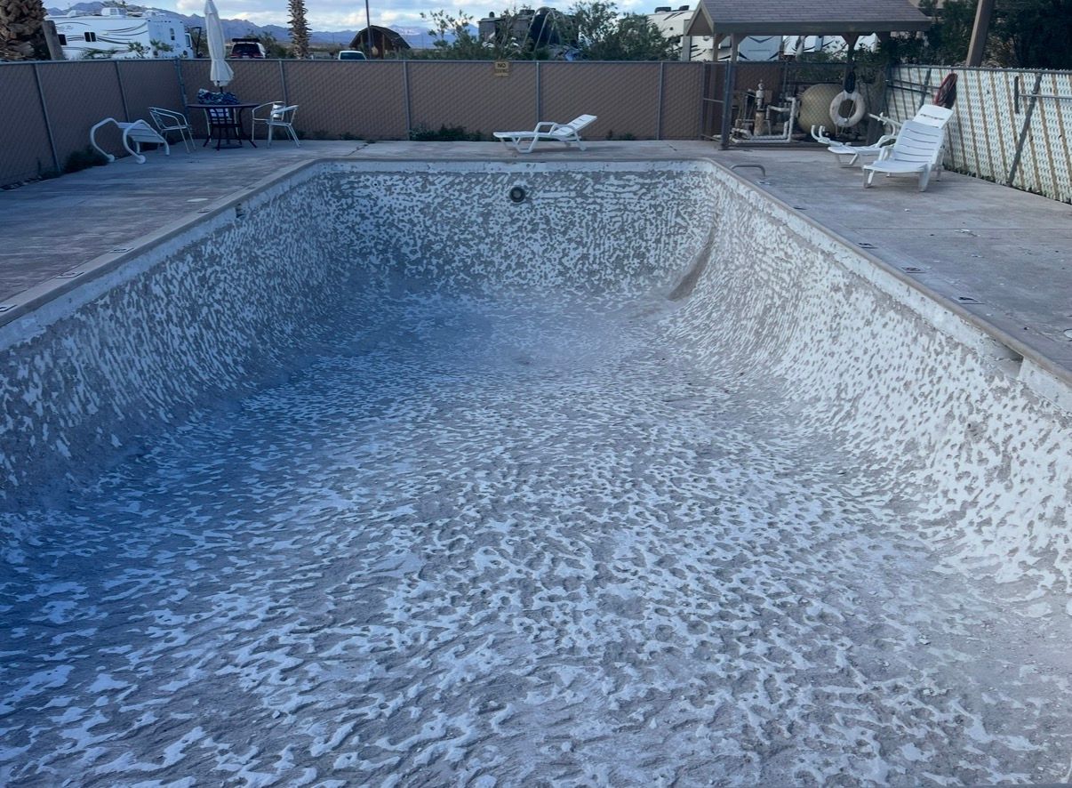 Empty swimming pool with a textured, whitish-blue interior and concrete surroundings. Fencing and houses in the background.