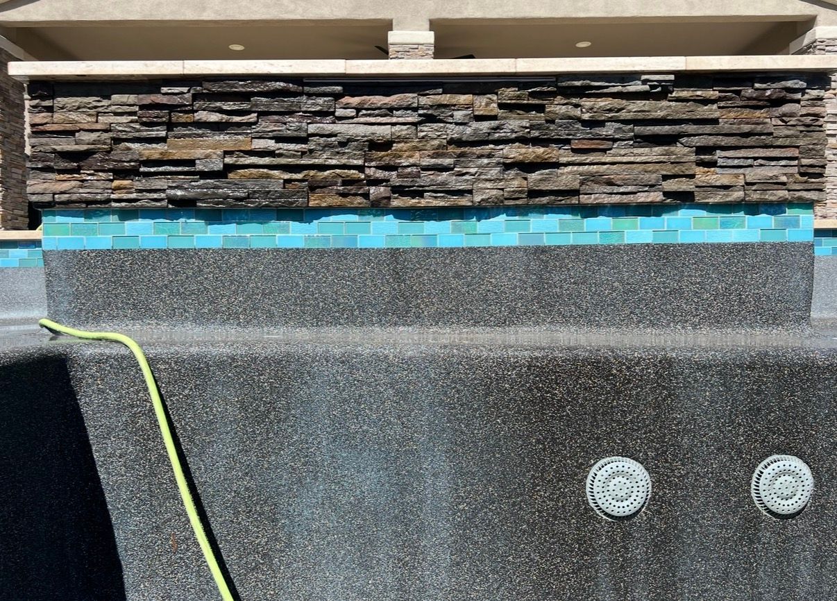 Pool interior with dark speckled plaster, blue tile, and stone veneer. A yellow hose rests nearby.