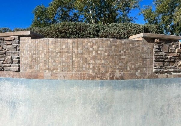 A stone wall water feature with tile backing and stone accents, topped with green bushes.