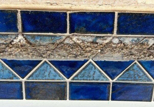 Close-up of a pool tile border with blue square and triangle tiles, showing crumbling grout.
