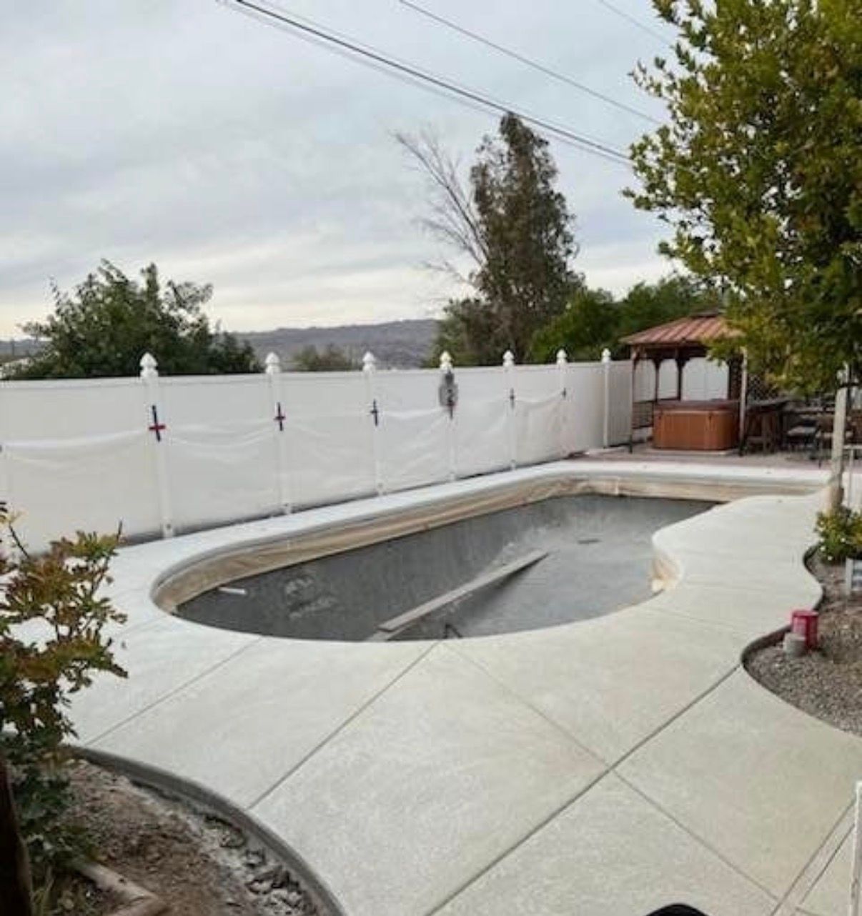 Concrete pool deck surrounds a covered pool, white fence, and a small gazebo in a backyard setting.